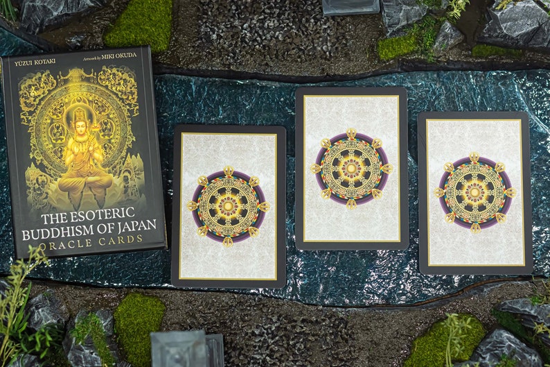 May include: A deck of "The Esoteric Buddhism of Japan Oracle Cards" by Yuzui Kotaki, with three cards showing a detailed wheel design. The book cover features a golden Buddha image. The cards are set on a textured surface.