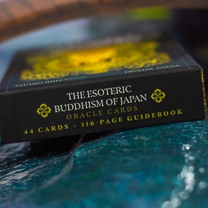 May include: A black box of "The Esoteric Buddhism of Japan Oracle Cards" with gold lettering and floral accents. The box states it contains 44 cards and a 116-page guidebook. The box is set against a blurred background.