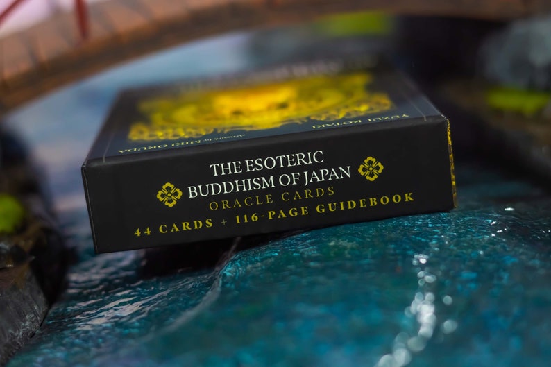 May include: A black box of "The Esoteric Buddhism of Japan Oracle Cards" with gold lettering and floral accents. The box states it contains 44 cards and a 116-page guidebook. The box is set against a blurred background.