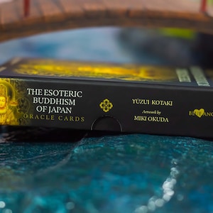 May include: A black box of "The Esoteric Buddhism of Japan Oracle Cards" with gold lettering and a gold illustration of a Buddha. The box is resting on a blue surface with a miniature bridge in the background. The text "Yuzui Kotaki" and "Artwork by Miki Okuda" are also visible.