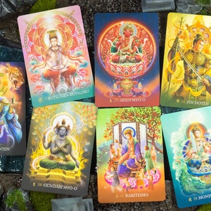 May include: A collection of colorful oracle cards featuring illustrations of spiritual figures. Each card displays a unique image with Japanese text and numbers. The cards are arranged on a surface with a water and rock-like background.