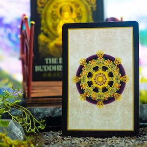 May include: A tarot card with a gold and purple wheel design, set against a neutral background. The card is framed in black. The image also features a bridge and a book with gold lettering, suggesting a spiritual or divination theme.