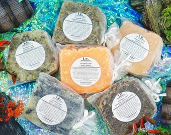 May include: Six bars of handmade soap wrapped in clear plastic bags with white ribbon ties. The soap bars are various colors, including green, brown, and orange. Each bar has a white label with the text "i.e. handcrafted soap" and the name of the scent. The scents are "Green Tea & Citrus", "Sweet Orange", "Lemongrass & Eucalyptus", "Chocolate, Coconut, & Coffee", "Lavender & Mint", and "Vanilla & Sandalwood".