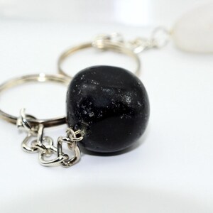 May include: A black, polished stone keychain with a silver-colored chain and ring. The stone is smooth and rounded, with a glossy finish. A white, teardrop-shaped stone is also visible in the background.