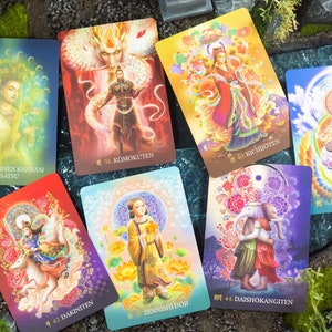 May include: A collection of colorful tarot cards featuring illustrations of deities and figures. Each card displays unique artwork with Japanese text and numbers. The cards are arranged on a natural background with rocks and greenery.