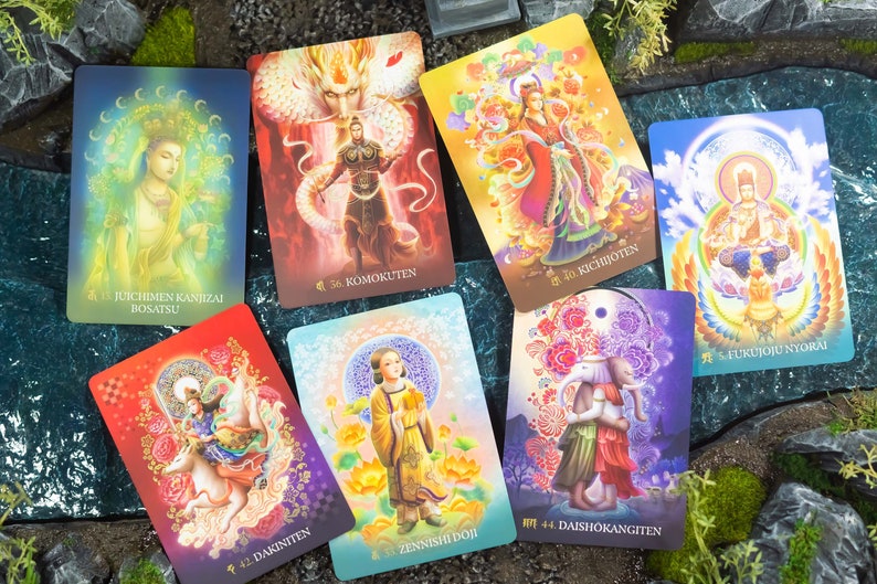 May include: A collection of colorful tarot cards featuring illustrations of deities and figures. Each card displays unique artwork with Japanese text and numbers. The cards are arranged on a natural background with rocks and greenery.