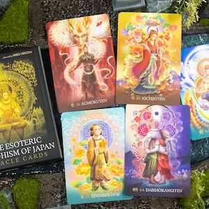 May include: A deck of "The Esoteric Buddhism of Japan Oracle Cards" with a black box featuring gold lettering and illustrations. Several colorful oracle cards are displayed, each with unique artwork and Japanese text. The cards depict various figures and symbols.