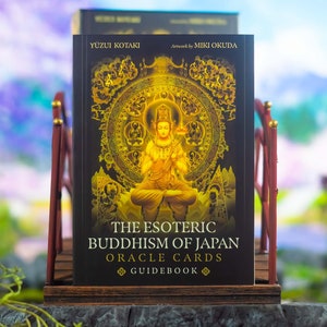 May include: A book titled "The Esoteric Buddhism of Japan Oracle Cards Guidebook" is displayed on a wooden stand. The cover features a golden illustration of a seated figure and intricate circular designs against a black background. The book is by Yuzui Kotaki, artwork by Miki Okuda.