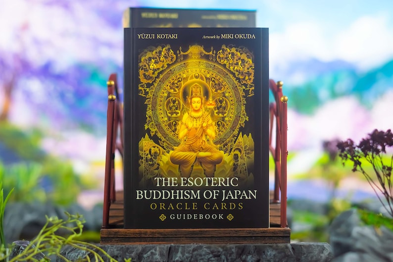 May include: A book titled "The Esoteric Buddhism of Japan Oracle Cards Guidebook" is displayed on a wooden stand. The cover features a golden illustration of a seated figure and intricate circular designs against a black background. The book is by Yuzui Kotaki, artwork by Miki Okuda.