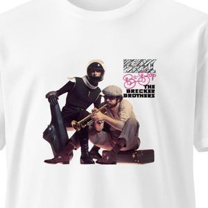 Op de afbeelding: Wit t-shirt met een afbeelding van twee mannen die muziekinstrumenten bespelen. De afbeelding bevat de tekst "The Brecker Brothers" en een fietsafbeelding in roze en zwart.