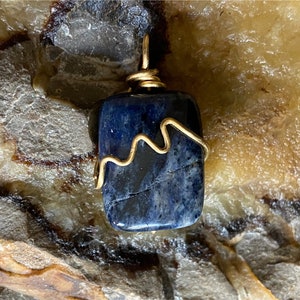 May include: A dark blue gemstone pendant with gold wire wrapping. The pendant is square shaped and has a rough, natural texture.