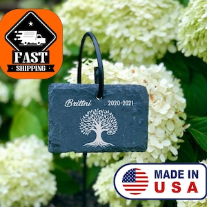 Könnte beinhalten: Schwarzer Schiefer-Gartenmarker mit einem Baumdesign und dem Text "Brittni 2020-2021". Der Marker ist an einem schwarzen Metallpfahl befestigt und von weißen Hortensienblüten umgeben.