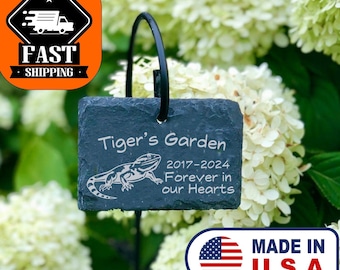 May include: Black metal garden stake with a slate stone marker that reads "Tiger's Garden 2017-2024 Forever in our Hearts". The stone marker features a lizard silhouette. The stake is in a bed of mulch with white hydrangeas in the background.