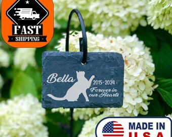 May include: Black slate garden marker with a cat silhouette and the text "Bella 2015-2024 Forever in our Hearts". The marker is attached to a black metal stake and is placed in the ground next to a white hydrangea bush.