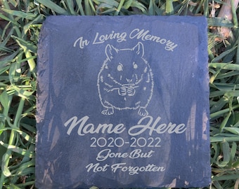 May include: A square, dark gray slate memorial stone with engraved text and a hamster illustration. The text reads "In Loving Memory," "Name Here," "2020-2022," and "Gone But Not Forgotten." The stone is set on a bed of green grass.