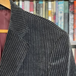 Könnte beinhalten: Ein schwarzer Cordblazer mit dünnen weißen Streifen. Der Blazer hat einen klassischen Stil mit einem Revers und einem Knopfverschluss.