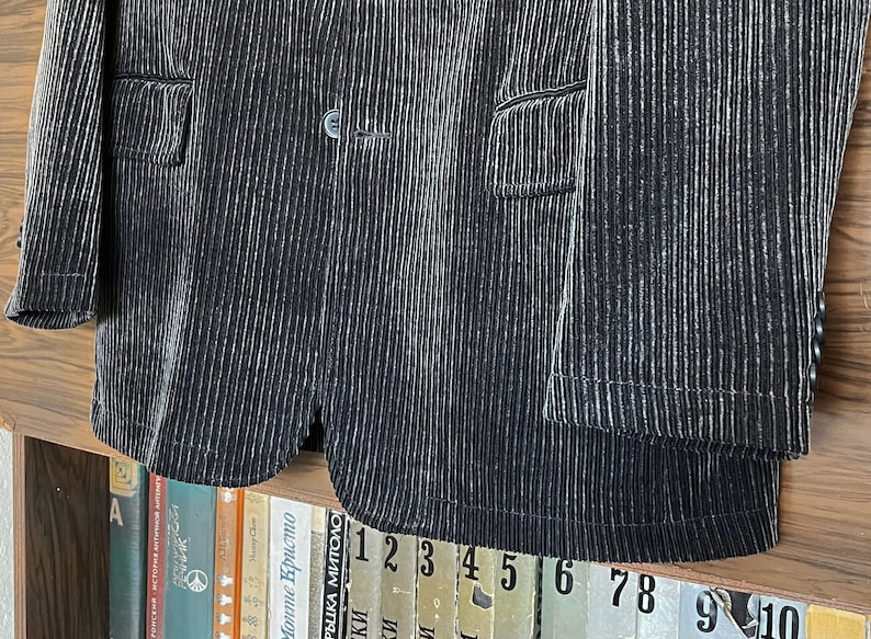 Może przedstawiać: Czarna marynarka z aksamitnego materiału w cienkie białe paski. Marynarka zapinana jest na jeden guzik i ma dwie kieszenie.