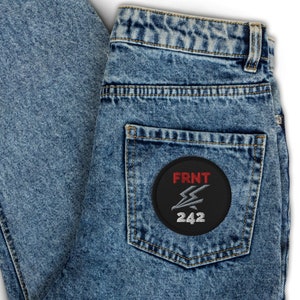Può includere: Un paio di jeans blu denim con una toppa nera sulla tasca posteriore. La toppa ha un fulmine rosso e il testo "FRNT 242" in bianco.