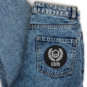 Peut inclure: Un jean bleu denim avec un patch noir sur la poche arrière. Le patch représente un crâne blanc avec un éclair et une couronne de laurier autour. Le texte "EBM" est écrit sous le crâne.