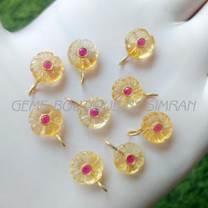 Könnte beinhalten: Eine Sammlung von blütenförmigen Edelsteinanhängern. Jeder Anhänger hat ein gelbes, durchscheinendes Blütenblattdesign mit einem kleinen, runden, roten Zentrum. Die Anhänger haben eine goldfarbene Schlaufe zum Aufhängen. Der Text "GEMS BOUTIQUE SIMRAN" ist sichtbar.