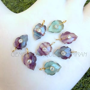 Peut inclure: Pendentifs en forme de feuille avec des bélières dorées. Les pendentifs sont en fluorite facettée multicolore, avec une pierre ronde et transparente sertie au centre de chaque feuille. Le texte "GEMS BOUTIQUE IMRAN" est visible.