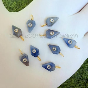 May include: A collection of blue gemstone pendants with gold-colored loops and small, clear accent stones. The pendants are in a geometric shape and are displayed on a white surface. The text "GL BOUTIQUE SIMRAN" is visible.