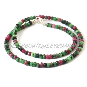 May include: A double-strand necklace featuring small, faceted beads in green, red, and black. The beads are strung together and secured with a silver clasp. The text "GEMSBoutiquebySimran" is visible. The necklace is a piece of jewellery.