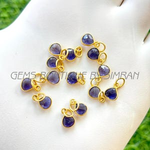 May include: A collection of gold-colored jewelry findings, each featuring a teardrop-shaped, deep blue gemstone. The gemstones are set in gold-colored metal with a small loop for attaching to other jewelry components. The text "GEMS BOUTIQUE BY SIMRAN" is visible.