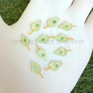 May include: A collection of light green gemstone pendants with a geometric shape, each featuring a small, round, green gem in the center and a gold-colored loop. The text "GEMS BOUTIQUE BY SIMRAN" is visible.