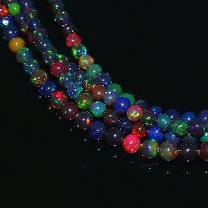 May include: Strands of iridescent opal beads in various colors, including blue, green, red, and yellow. The beads are round and have a polished finish, reflecting light. The beads are strung together, creating a necklace or bracelet.