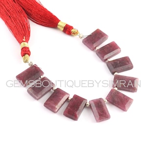 Puede incluir: Una hebra de cuentas de rubí rojo con un borla roja en cada extremo. Las cuentas son de forma rectangular y tienen un acabado pulido. Las cuentas están enhebradas en un alambre de plata.
