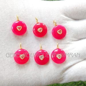 May include: Six circular, hot pink pendants with gold-coloured loops. Each pendant has a heart-shaped, clear stone in the centre. The text "GEMS BOUTIQUE BY SIMRAN" is visible in the image.