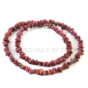 Puede incluir: Un collar hecho de pequeñas piedras preciosas de color rosa rojizo de forma irregular. Las piedras están ensartadas, formando un bucle continuo. El collar tiene un cierre plateado. El fondo es blanco.