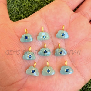 Puede incluir: Nueve colgantes en forma de nube con lazos dorados. Cada colgante es de color azul verdoso claro con una piedra circular azul oscuro en el centro. El texto "GEMS BOUTIQUE BY IMRAN" es visible.