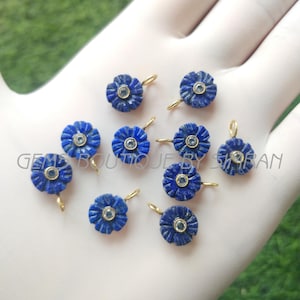 Może przedstawiać: Kolekcja niebieskich zawieszek w kształcie kwiatów ze złotymi pętelkami. Każda zawieszka ma centralny, jasnoniebieski kamień. Zawieszki są ułożone na białej powierzchni z zielonym tłem. Widoczny jest tekst "GEMS BOUTIQUE BY SHABAN".