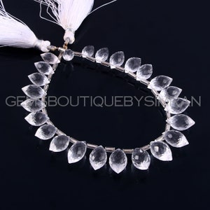 Puede incluir: Un collar de piedras preciosas transparentes en forma de lágrima facetadas, ensartadas con pequeñas cuentas plateadas. Las piedras están dispuestas simétricamente. Hay borlas blancas en la parte superior. El texto "GEMSBoutiquebySiman" es visible.