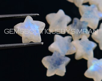 May include: A group of white moonstone star-shaped cabochons with iridescent blue and white flashes. The stones are scattered on a black surface. One star is held in tweezers.