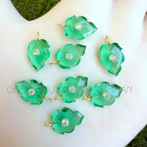 Peut inclure: Sept pendentifs verts en forme de feuille avec des bélières dorées. Chaque pendentif présente un design facetté et une petite pierre précieuse ronde et transparente au centre. Les pendentifs sont présentés sur une surface claire.