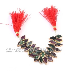 Puede incluir: Collar con piedras preciosas en forma de rombo en tonos verdes, morados y negros, con bordes dorados. Dos borlas rojas adornan la parte superior. Las piedras están dispuestas en un patrón curvo. Adecuado para diversas ocasiones.