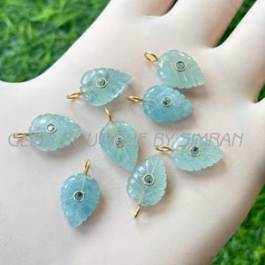 Puede incluir: Ocho colgantes de hojas de aguamarina con aros dorados y pequeñas gemas azules en el centro. Los colgantes se sostienen en una mano enguantada sobre un fondo verde. El texto "GEMS BOUTIQUE BY SIMRAN" es visible.