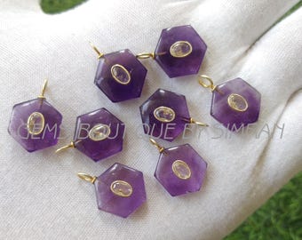 May include: A collection of amethyst gemstone pendants with gold-colored bails. Each hexagonal pendant features a central oval-shaped, light-colored stone. The pendants are displayed on a white surface with a green background.