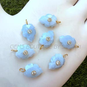 Op de afbeelding: Zeven lichtblauwe edelsteenhangers in de vorm van een blad, elk met een kleine, ronde, heldere steen in het midden. De hangers hebben goudkleurige lussen om op te hangen. De tekst "GEMS BOUTIQUE BY SIMRAN" is zichtbaar.