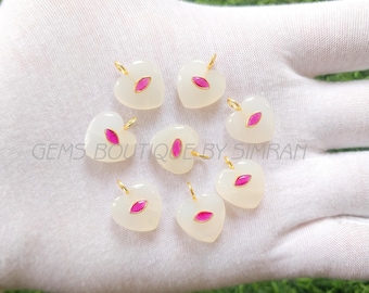 May include: Seven heart-shaped pendants with a gold bail. Each pendant is white with a pink center and a gold border. The pendants are arranged on a white surface. The text "GEMS BOUTIQUE BY SIMRAN" is visible.