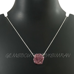 May include: A silver chain necklace with a raw pink ruby pendant. The pendant is a natural, rough-cut stone with a deep pink color. The necklace is a simple, delicate design that would make a beautiful gift.
