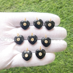 Puede incluir: Ocho colgantes en forma de corazón de color negro, cada uno con una argolla dorada y una piedra redonda transparente en el centro. Los colgantes se muestran sobre una superficie blanca, sobre un fondo verde.