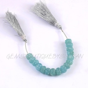 May include: A strand of light blue aquamarine beads with a carved pumpkin shape. The beads are strung on a silver wire with silver tassels at each end.