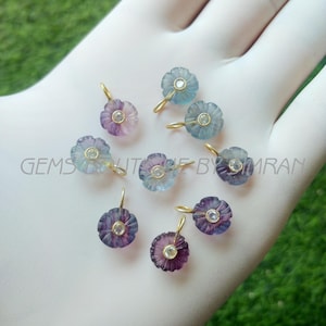May include: A collection of flower-shaped gemstone pendants in shades of purple, blue, and clear. Each pendant has a gold-colored loop and a small, round, clear stone in the center. The pendants are displayed on a white surface.