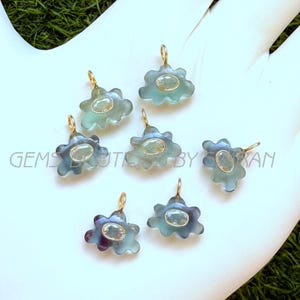 Op de afbeelding: Zeven wolkvormige hangers van doorschijnend blauwe en paarse fluoriet met een centrale ovale edelsteen en een goudkleurig metalen oogje. De tekst GEMS BOUTIQUE BY KIRAN is zichtbaar in het midden van de opstelling van deze sieraden.