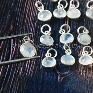 Op de afbeelding: Een verzameling zilveren bedels met ronde, witte maansteen edelstenen. De bedels zijn aan een zilveren ketting geregen en zijn perfect om een ​​toets van elegantie toe te voegen aan elk sieraad.