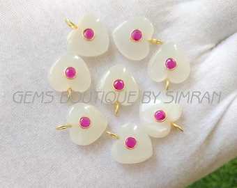May include: Heart-shaped pendants with a central pink gemstone and gold-colored loops. The pendants are white and arranged in a circular pattern. The text "GEMS BOUTIQUE BY SIMRAN" is visible.